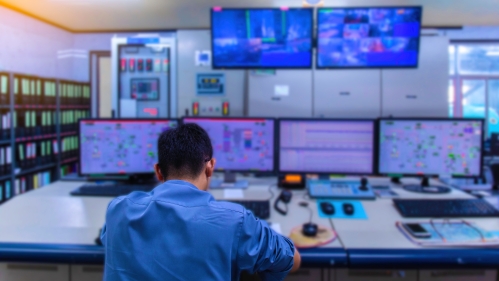 The back of a man sitting at a desk with four computer monitors in front of him and two television monitors hanging from the ceiling.