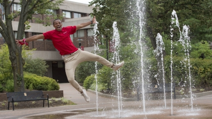 Male student jumps in the air in front a fountain sprsying water. He's wearing khaki pants and red Rutgers shirt.