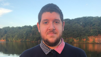 Head shot of male wearing a dark blue sweater with red, white and blue stars and striped collar standing with a lake in the background. 