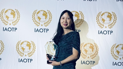 Asian female with long black hair standing in front of an IAOTP step and repeat banner in a black long gown holding an award. made of glass
