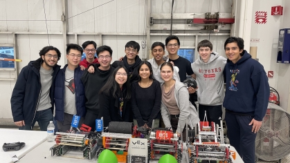 Twelve college students in a lab posing behind several robots on a table.