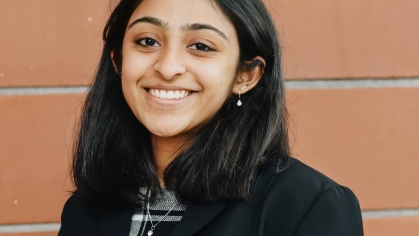 Young woman poses in front of a brick wall. She has black hair and is wearing a black jacket.