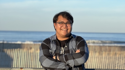 Male student stands wearing a black and grey plaid button down shirt, black horn rimmed eyeglasses with his arms folded with the ocean in the background. 