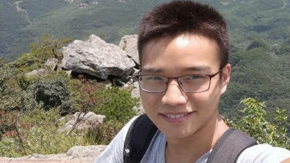 Student with short black hair wearing eyeglasses standing on a mountain with the valley in the background.  