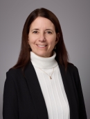 Headshot of female with long brown hair wearing a white turtleneck sweater, a gold necklace, and a black suit jacket.