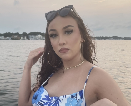 Woman poses along the shoreline. She has dark hair, sunglasses are perched on her head. She is wearing a blue and white top.