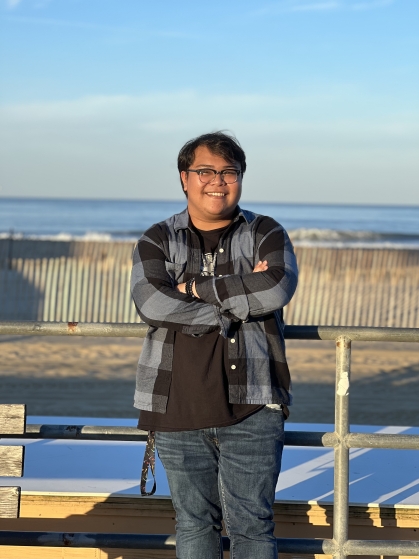 Male student stands wearing a black and grey plaid button down shirt, black horn rimmed eyeglasses with his arms folded with the ocean in the background. 