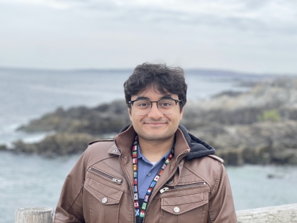 Male engineering student headshot posing outdoors with water and a rocky coastline behind him. He has dark hair, glasses and is wearing a brown jacket over a blue shirt.