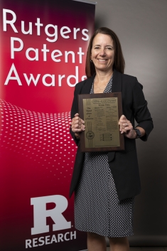 Woman wearing a black and white dress and black blazer holds award plaque in front of a red banner that says Rutgers Patent Awards.of a